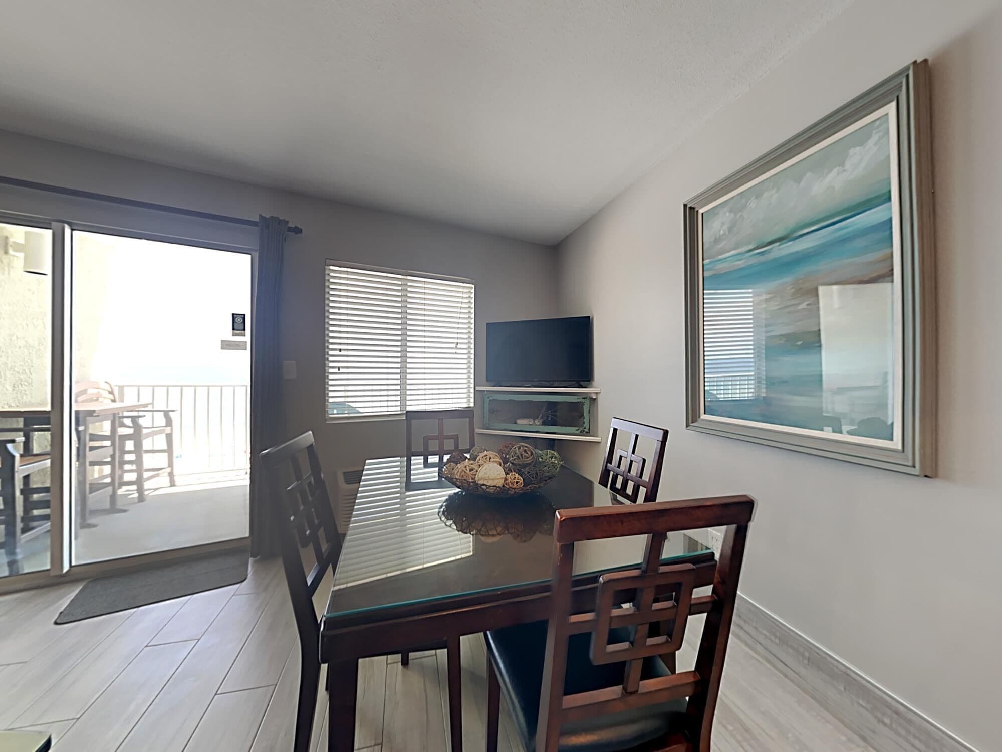 A dining area with a glass-top table, wooden chairs, a television on a shelf, and a painting on the wall.