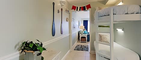 A narrow hallway with a bunk bed on the right, a potted plant on the left, and a door leading to a room with a blue curtain.