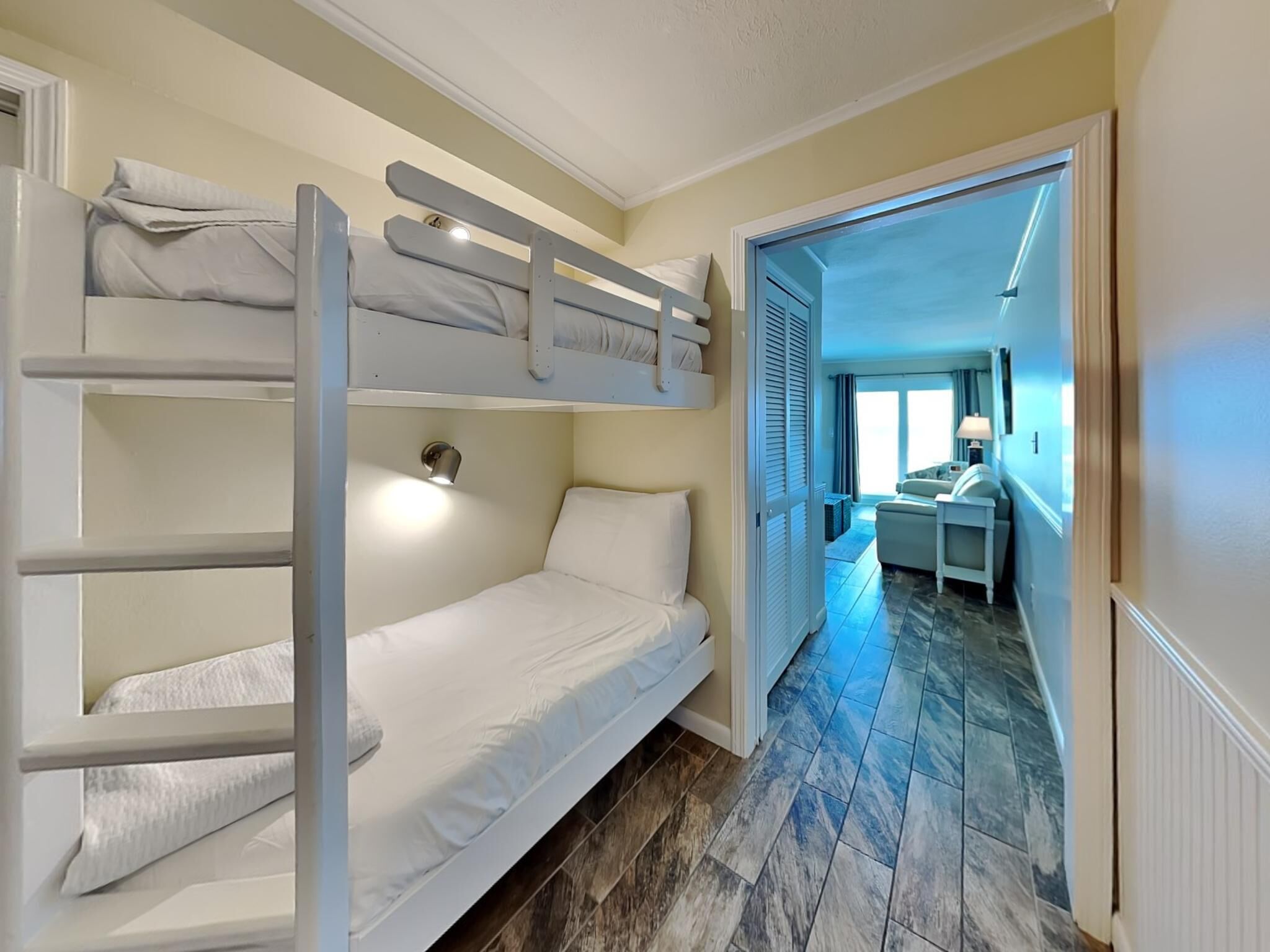 A bunk bed room with a small desk and a chair, a window with blinds, and a hallway leading to a living area.