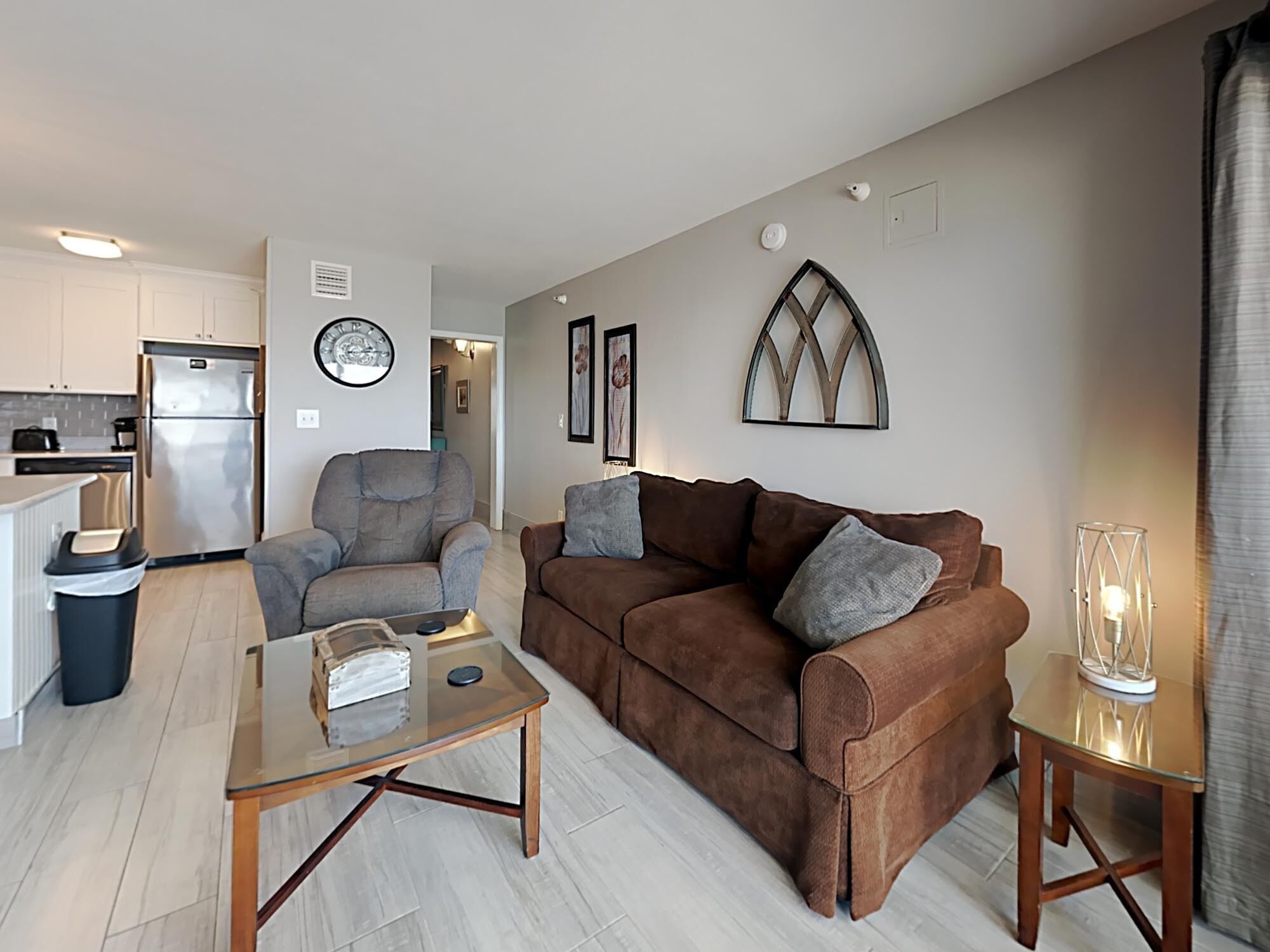 A modern living room with a brown sofa, a glass coffee table, and a kitchen in the background.