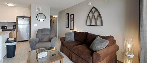 A modern living room with a brown sofa, a glass coffee table, and a kitchen in the background.