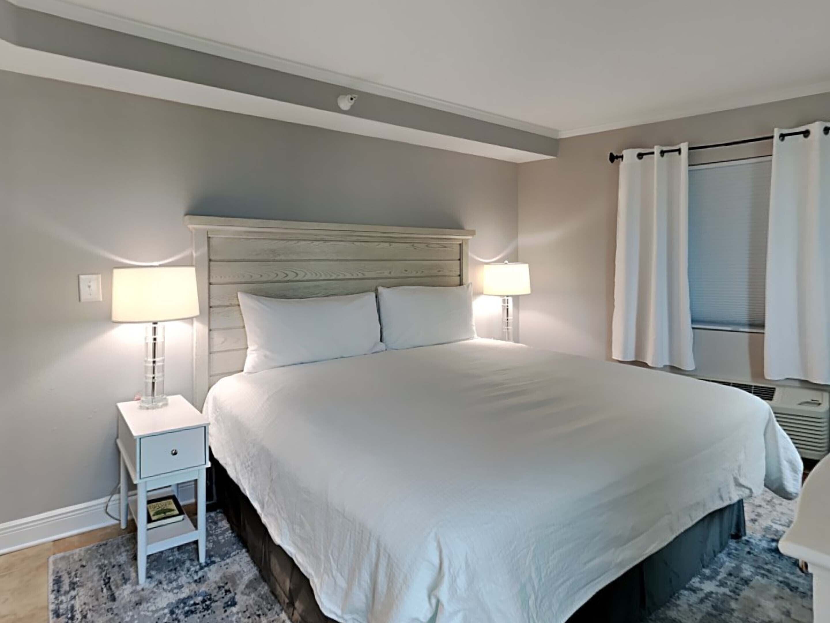 A bedroom with a large bed, two bedside tables with lamps, a ceiling-mounted security camera, and a window with curtains.