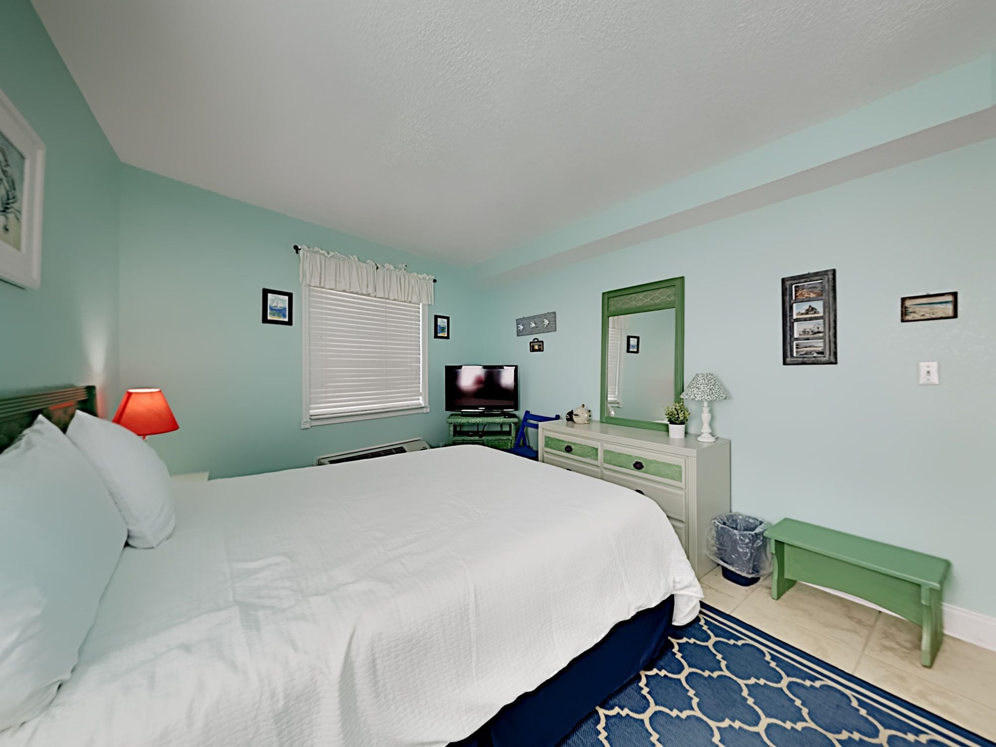 A bedroom with a bed, a television, a dresser, a mirror, and a window with blinds.
