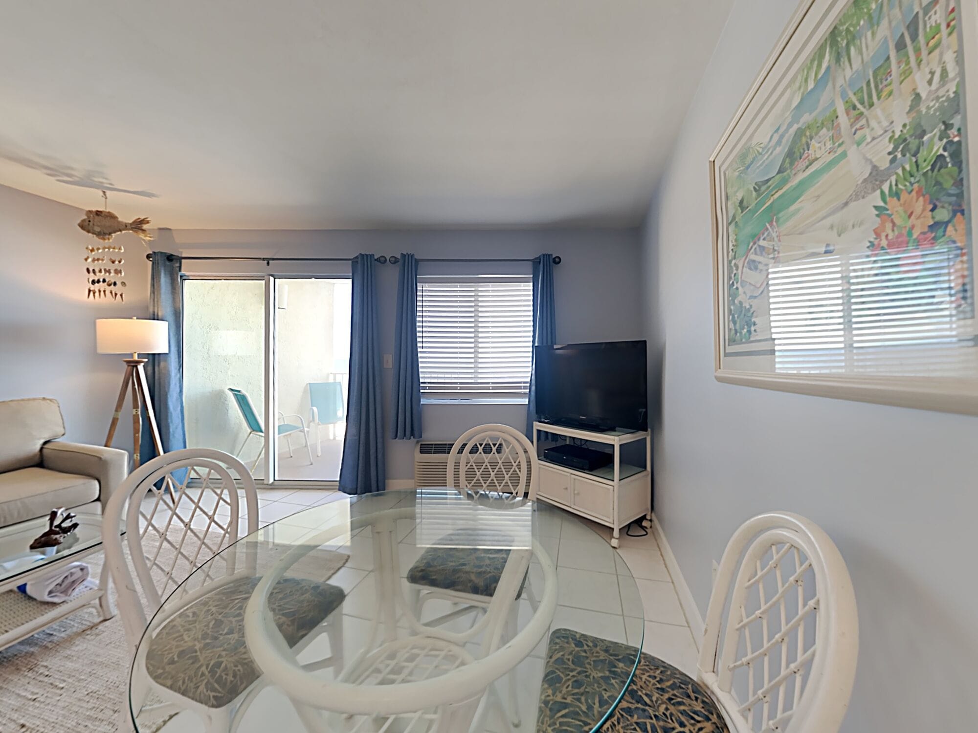 A living room with a glass-top dining table, white chairs, a sofa, a TV on a white cabinet, and a large window with blue curtains.