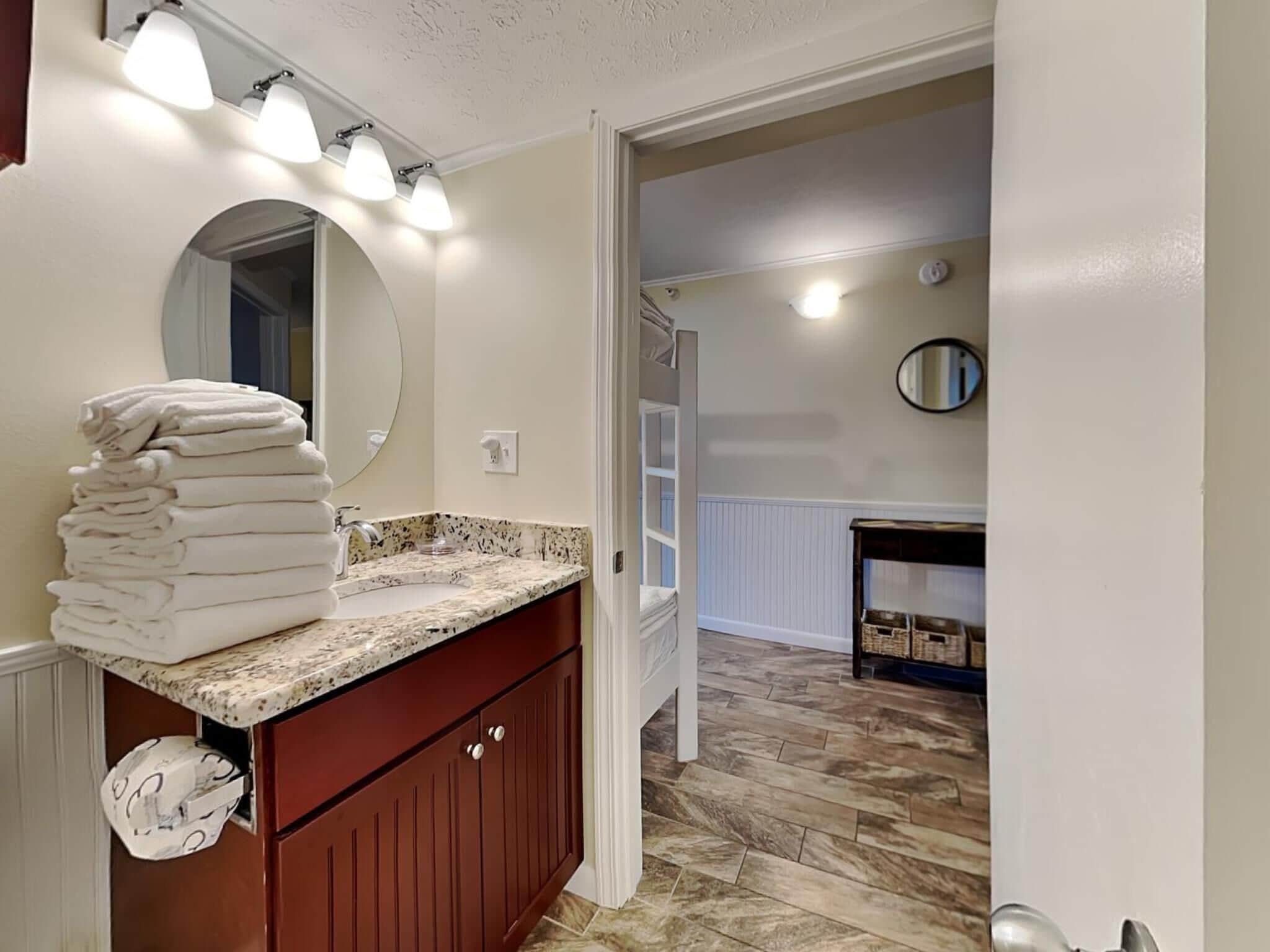 A bathroom with a marble countertop, a stack of white towels, a round mirror, and a door leading to another room.