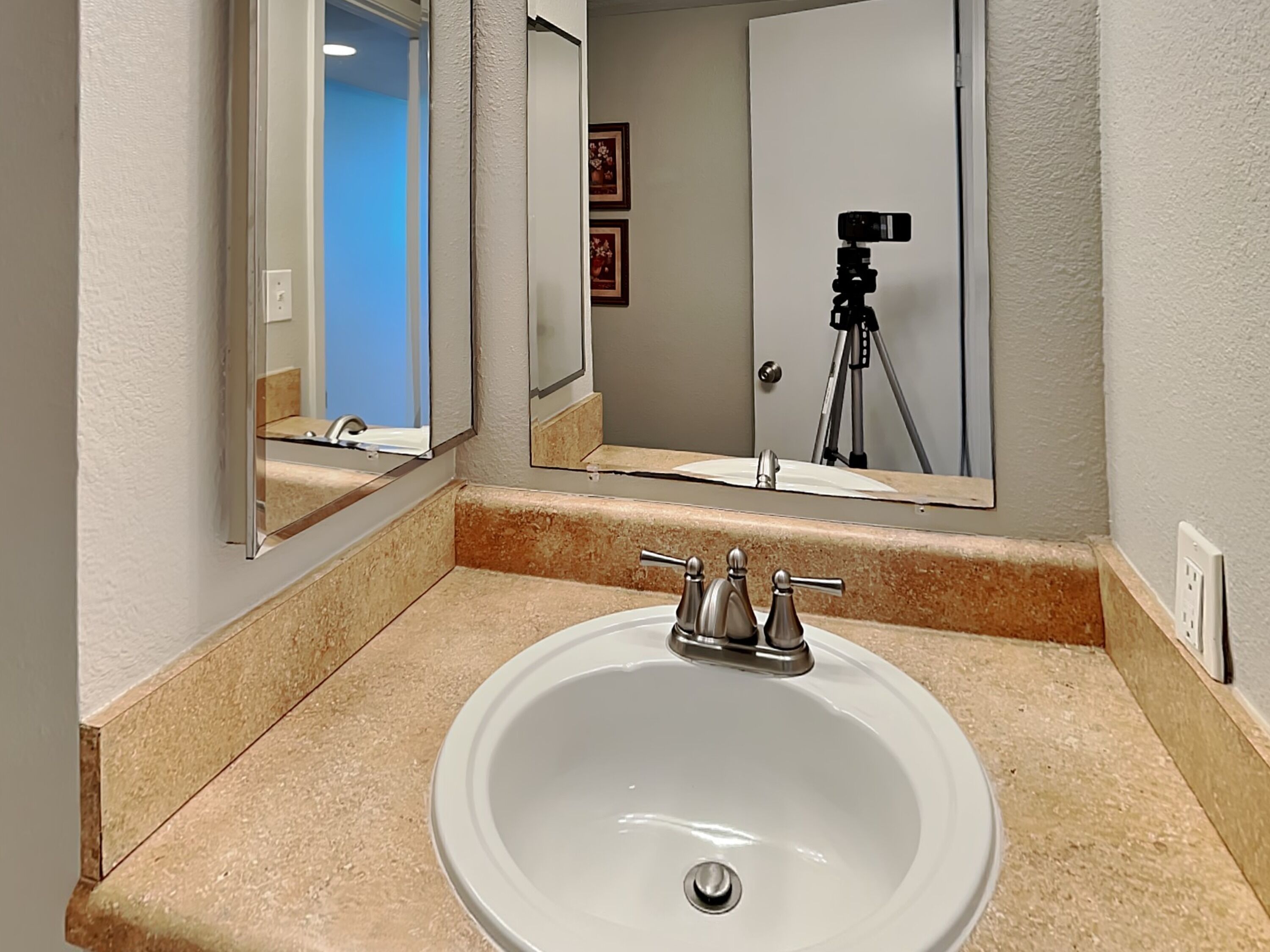 Une salle de bain avec un lavabo blanc, un miroir et un appareil photo posé sur un trépied à l’arrière-plan.