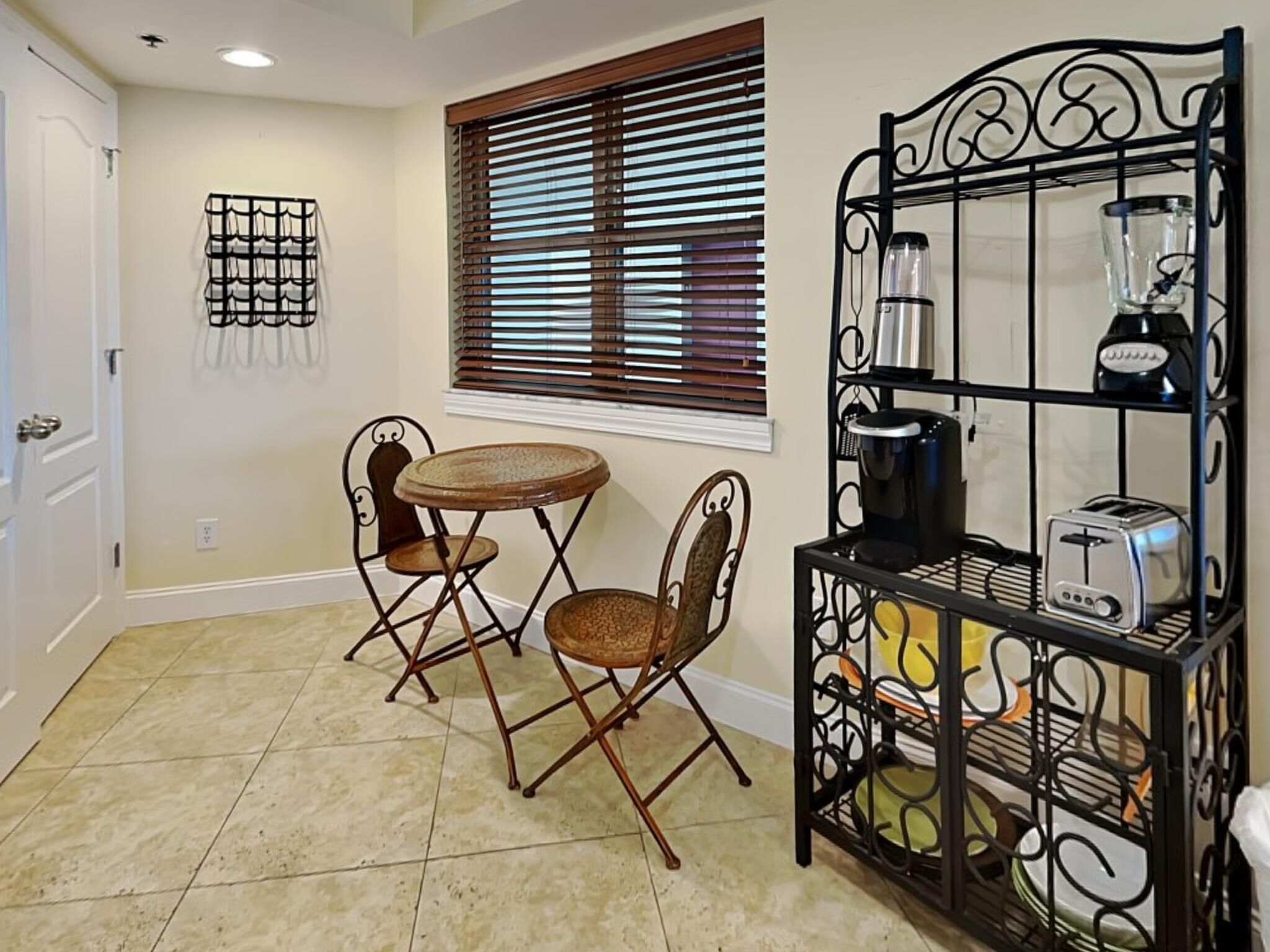 A kitchen with a dining area, including a small round table and two chairs, a window with blinds, and a black metal rack with kitchen appliances.