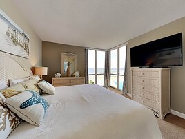 A bedroom with a large bed, a dresser, a television, and a view of the ocean.