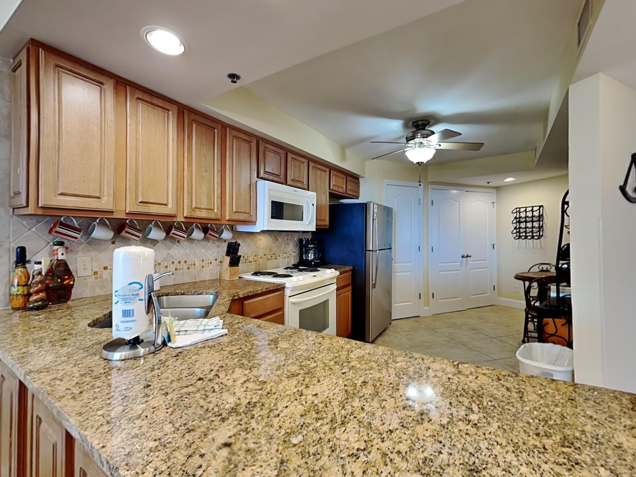 A kitchen with wooden cabinets, a granite countertop, a microwave, a refrigerator, and a ceiling fan.