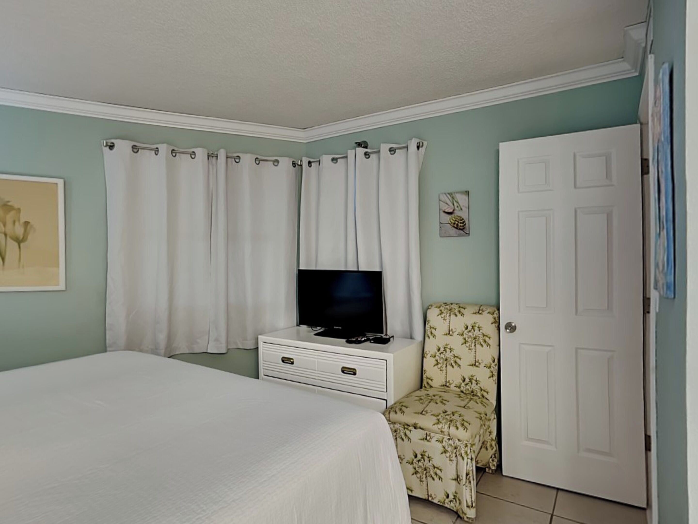 Une chambre à coucher avec un lit, un téléviseur posé sur une commode, une chaise et une porte.