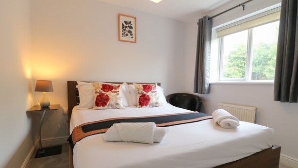 A neatly made bed with white linens and floral pillows, a bedside lamp, and a view of greenery through a window.