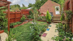 A brick house with a wooden deck, a garden with various plants, and a concrete pathway.