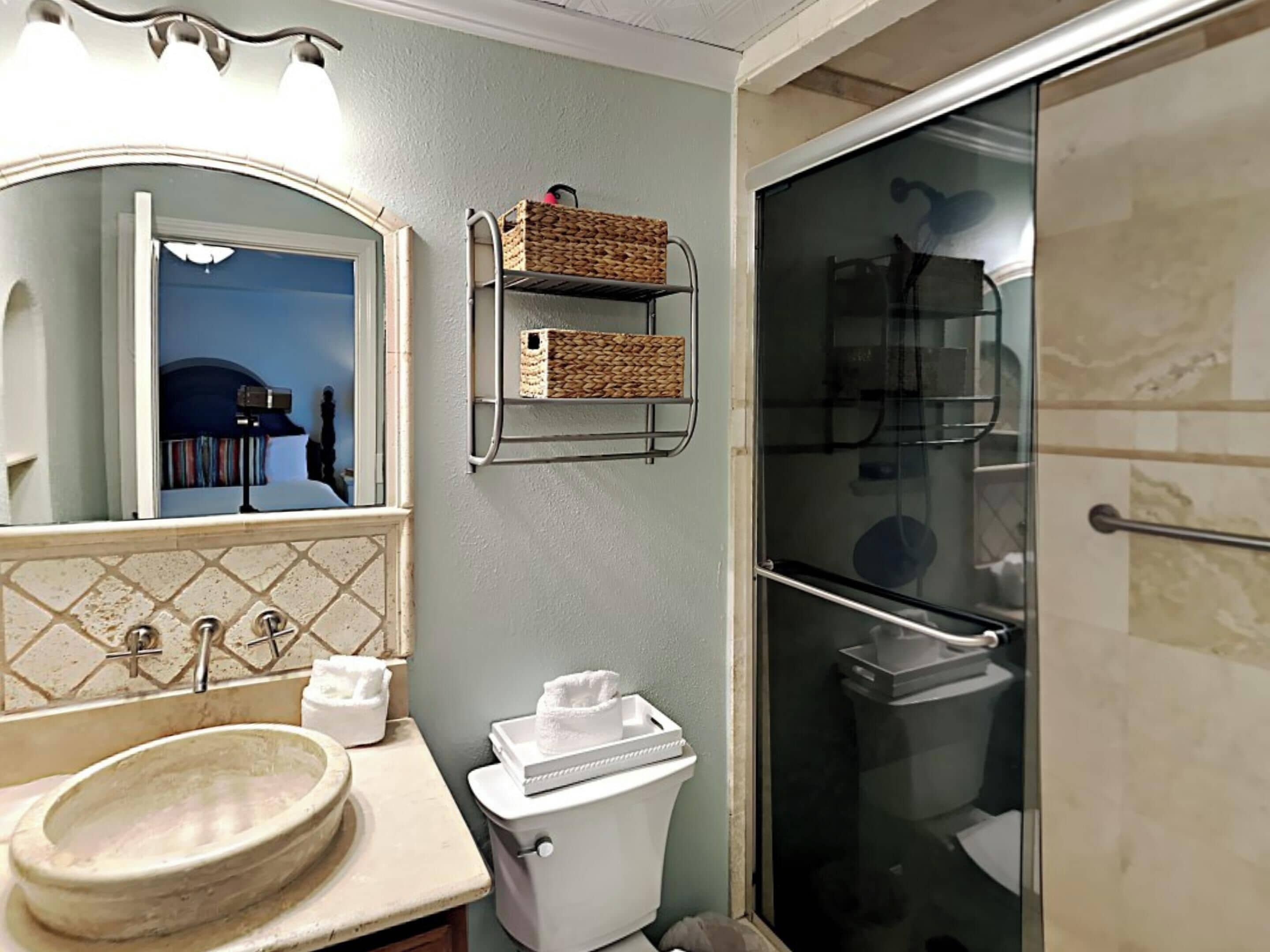 Un baño con lavamanos, inodoro y cabina de ducha de vidrio. Hay cestos de mimbre en un estante metálico sobre el inodoro.