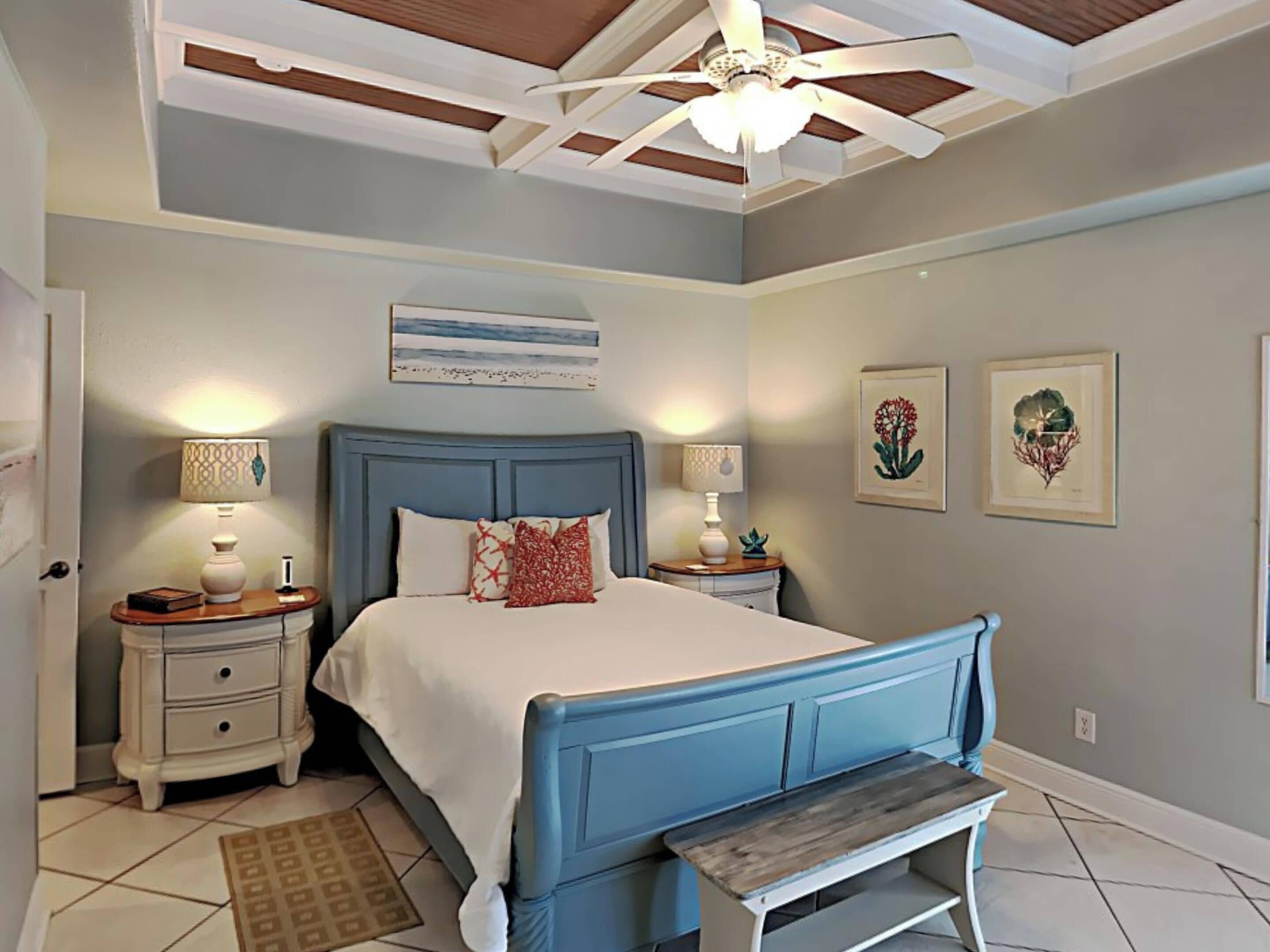 A bedroom with a bed, bedside tables, a bench, a ceiling fan, and artwork on the walls.