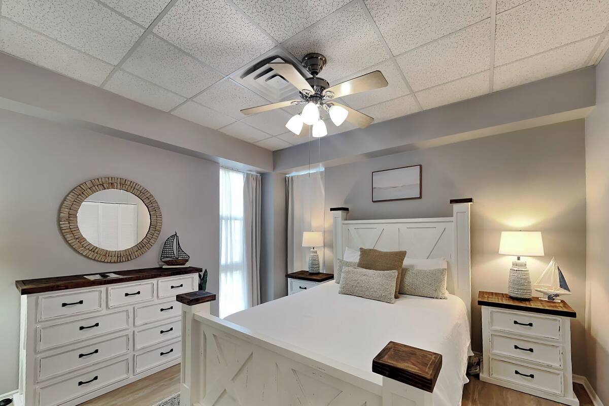 A bedroom with a bed, nightstands, a dresser, a ceiling fan, and a window with curtains.