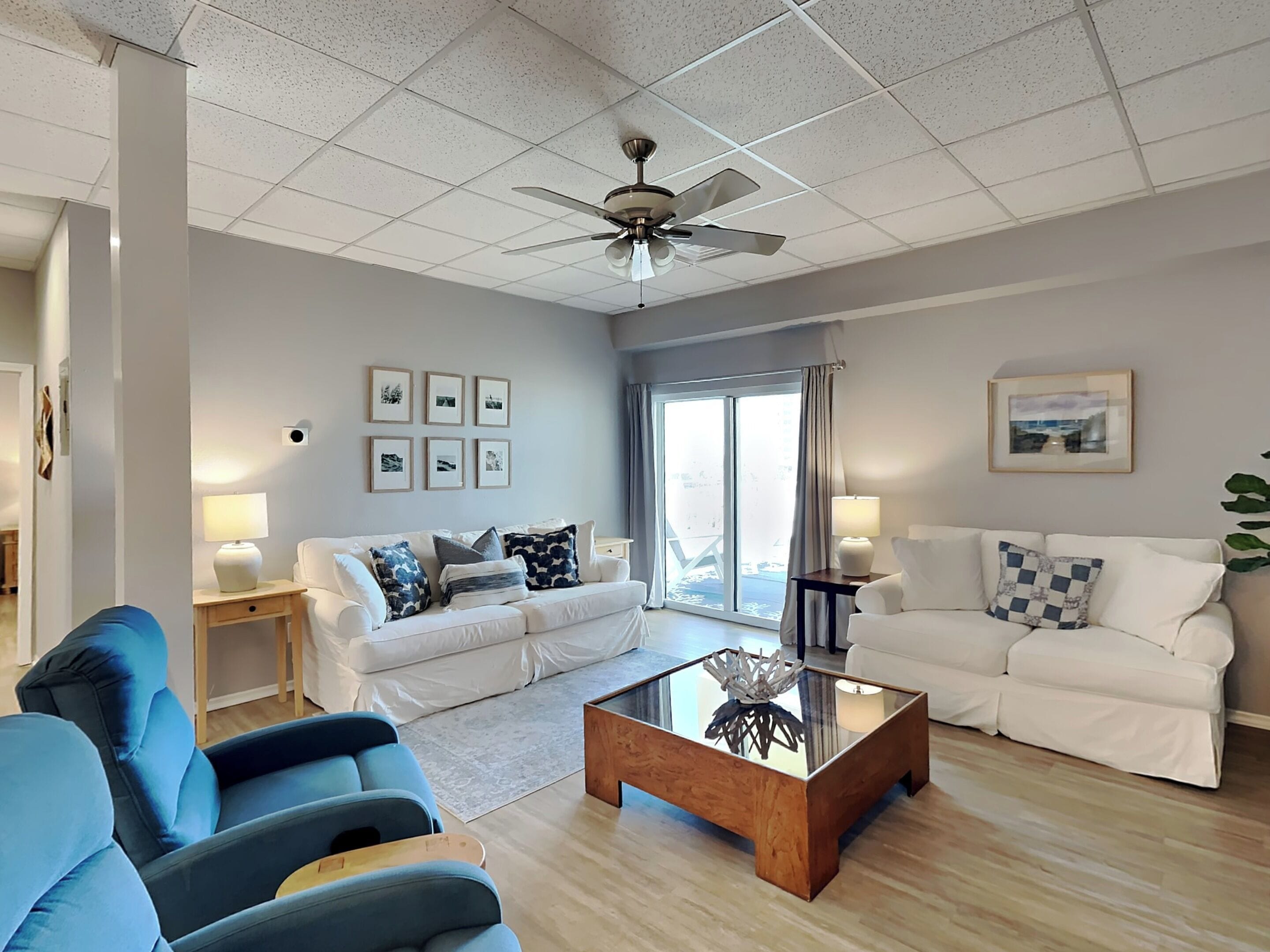 A spacious living room with a ceiling fan, a large window with curtains, and a white sofa with blue cushions.