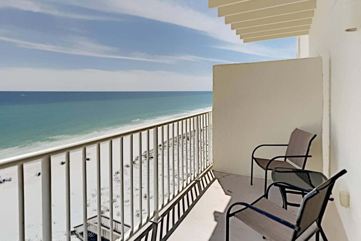 A balcony with a view of the beach and ocean. There are two chairs and a table.
