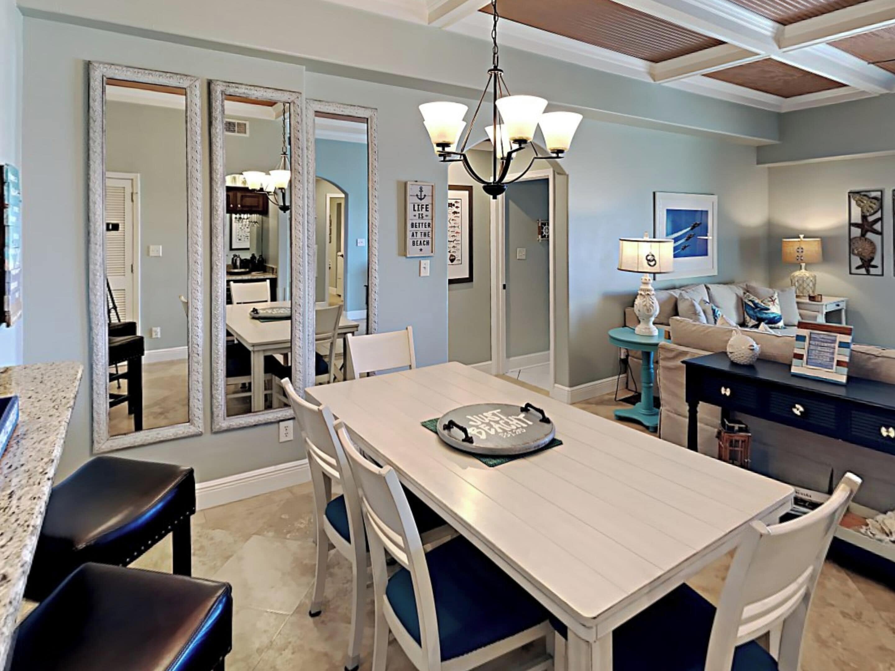 A dining area with a white table and chairs, a chandelier, and a kitchen in the background.