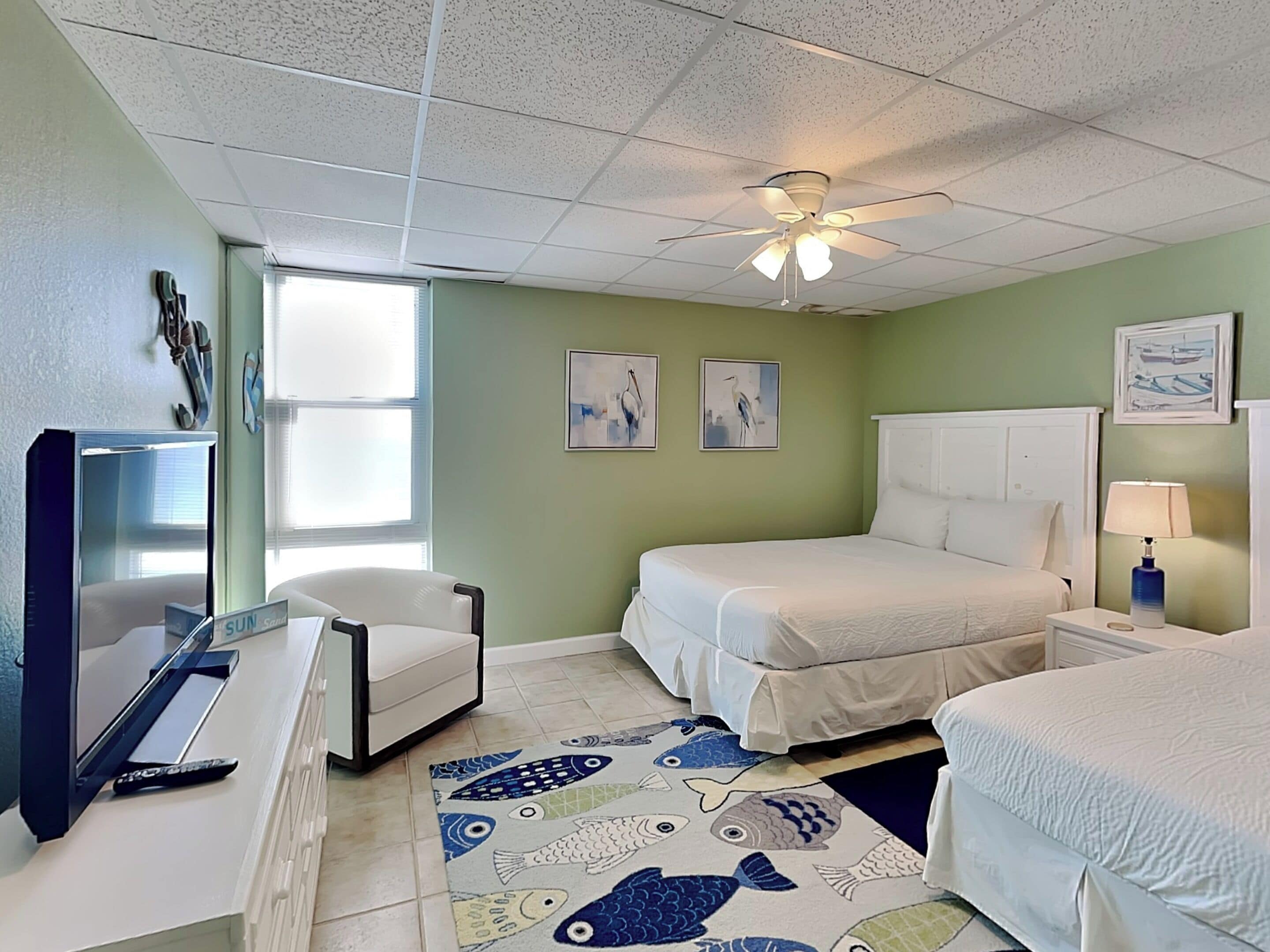 A hotel room with a bed, a desk, a chair, a ceiling fan, and artwork on the walls.