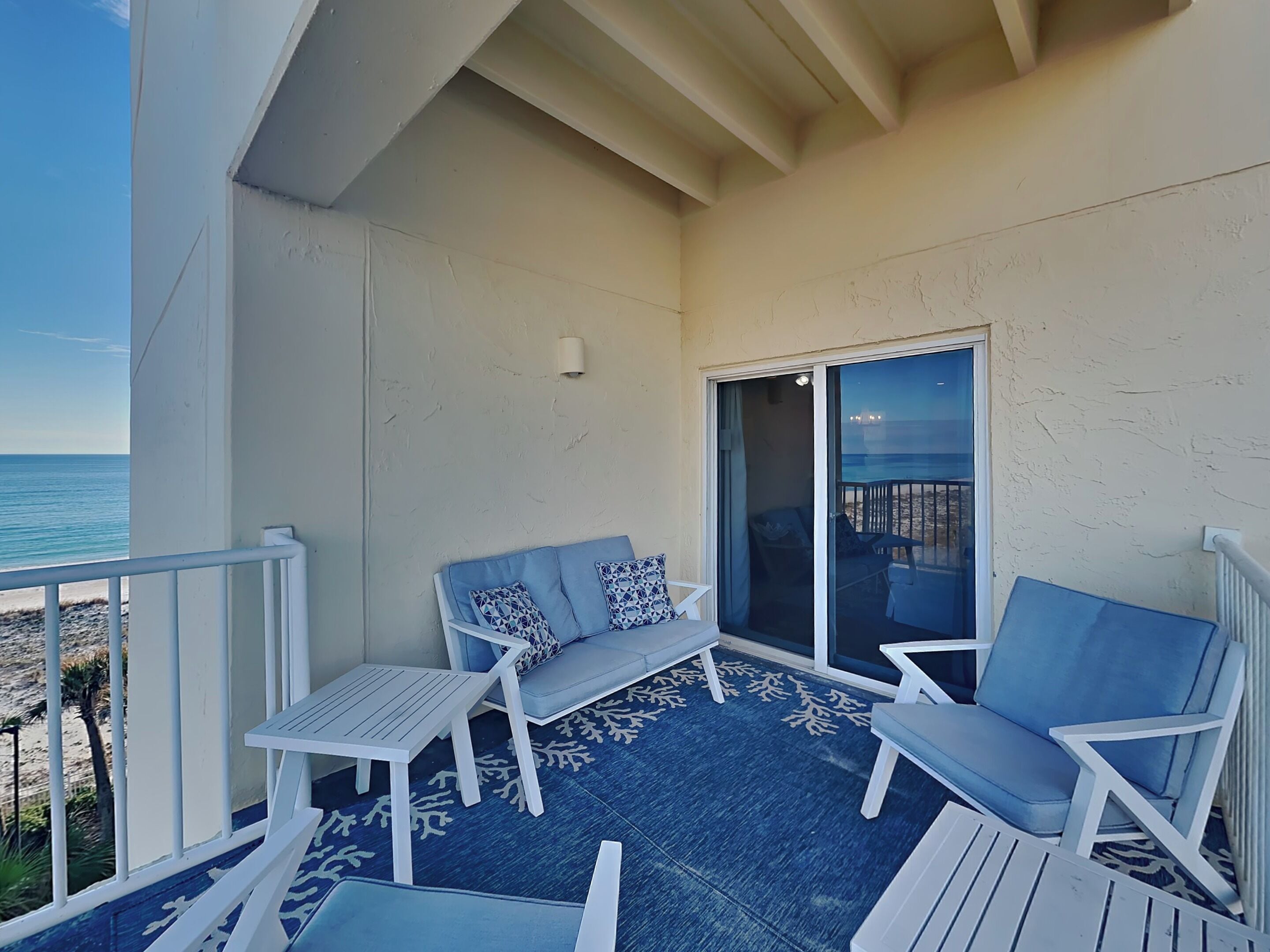 A balcony with a blue patterned rug, white furniture, and a view of the beach.