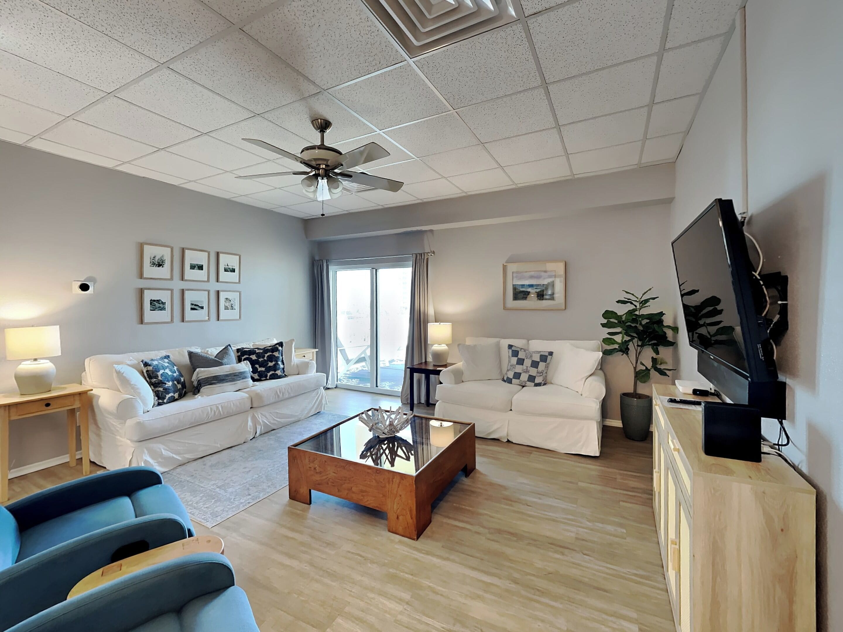 Una sala moderna con un sofá blanco, sillones azules, una mesa de centro de madera y un ventilador de techo.
