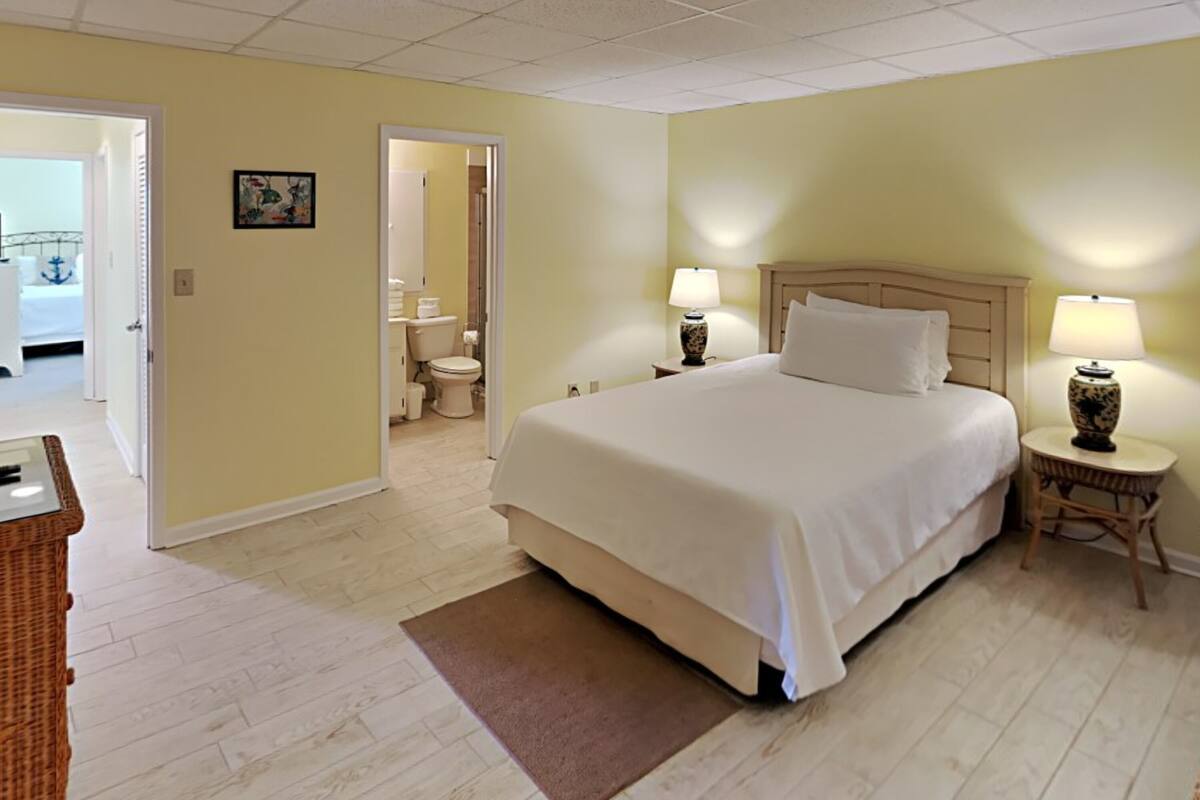 A hotel room with a large bed, two bedside lamps, a small table, and a door leading to a bathroom.