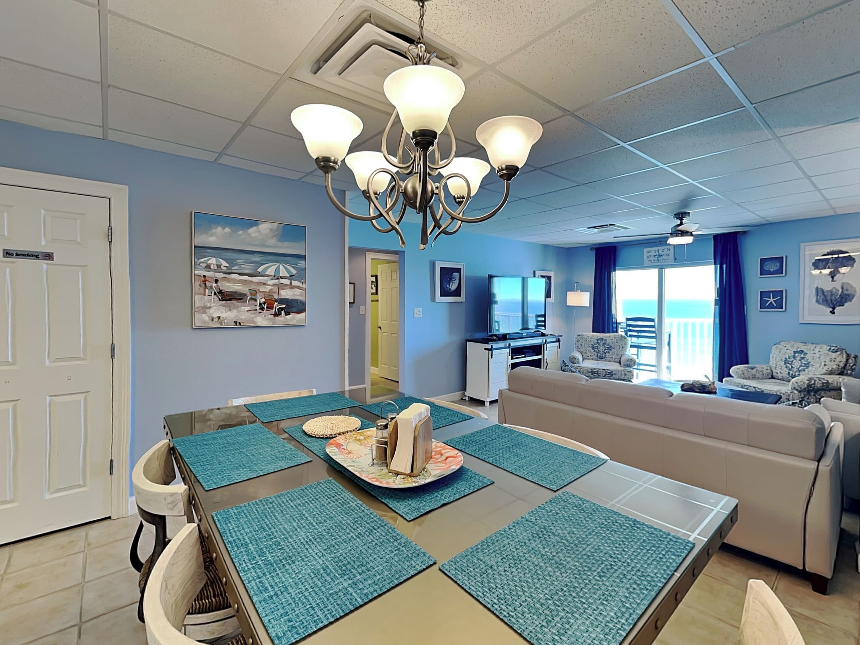 Un salon moderne avec un coin repas, agrémenté d’un lustre, de murs bleus, d’un tableau sur le thème de la plage et offrant une vue sur l’océan par le biais d’une grande fenêtre.