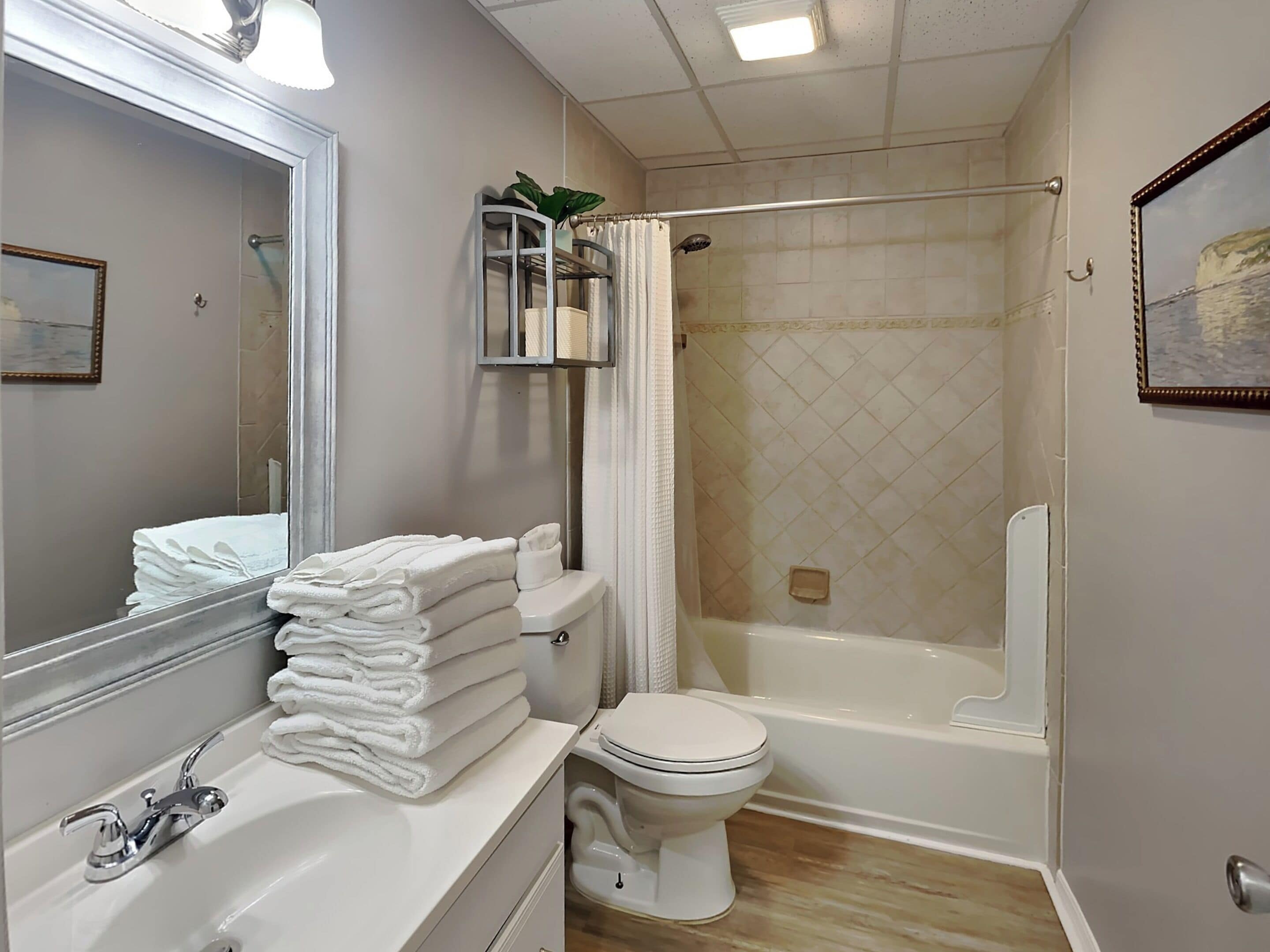 A bathroom with a sink, toilet, and bathtub. There is a towel rack with folded towels, a mirror, and a framed picture on the wall.
