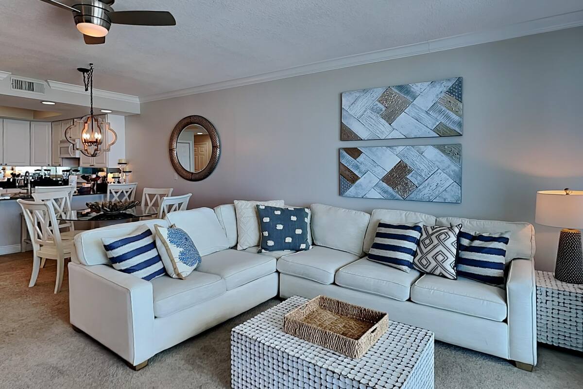 A living room with a white sectional sofa, a dining area with white chairs, a kitchen in the background, and two abstract wall art pieces.