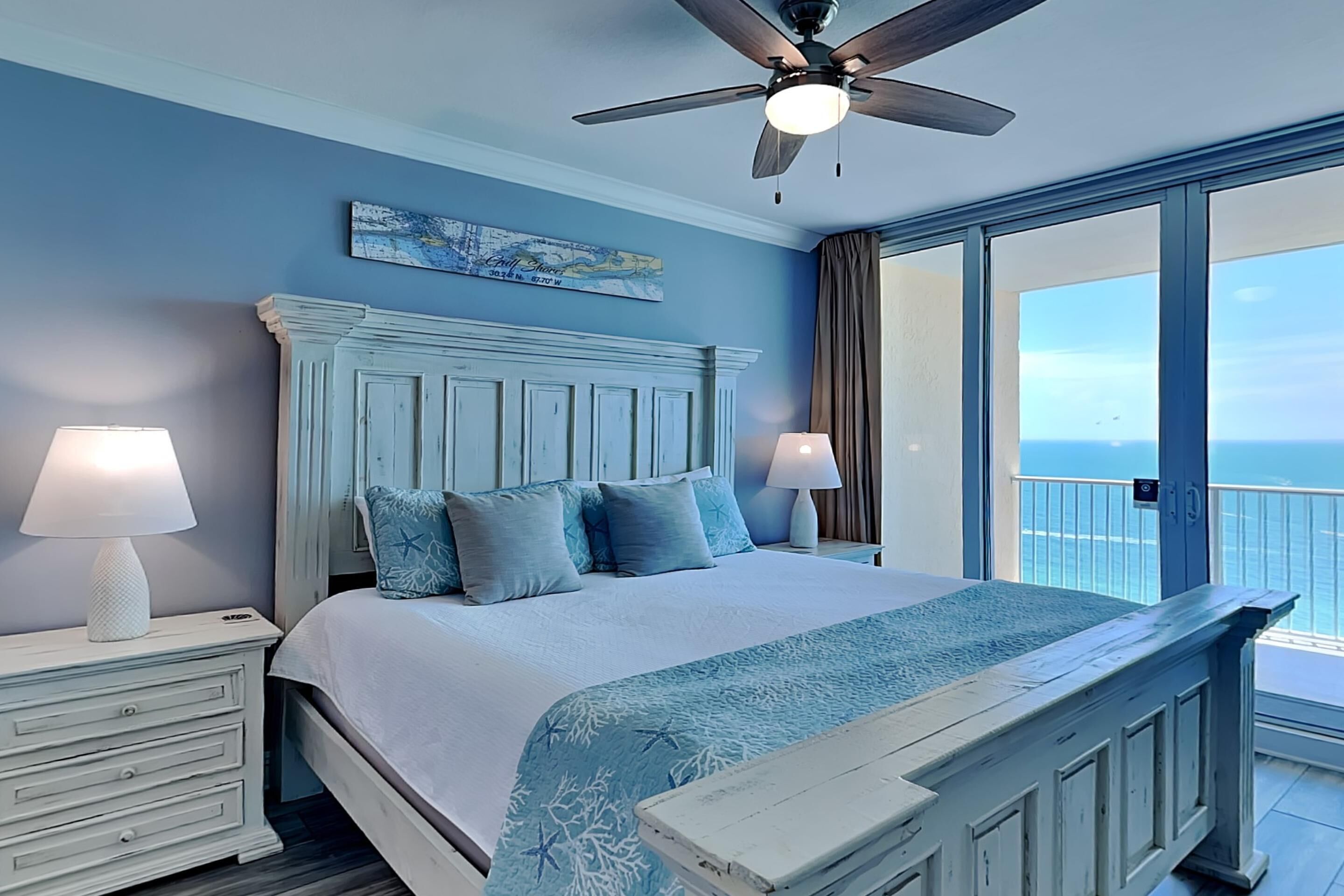 A bedroom with a large bed, two bedside tables with lamps, a ceiling fan, and a view of the ocean through a large window.