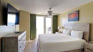 Quarto de hotel com cama, televisão, ventoinha no teto, um quadro e vista para o exterior.