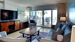 A modern living room with a flat-screen TV, a sofa, a coffee table, and a view of the outdoors through large sliding doors. - Sandy Key by Southern Vacation Rentals (Pensacola)