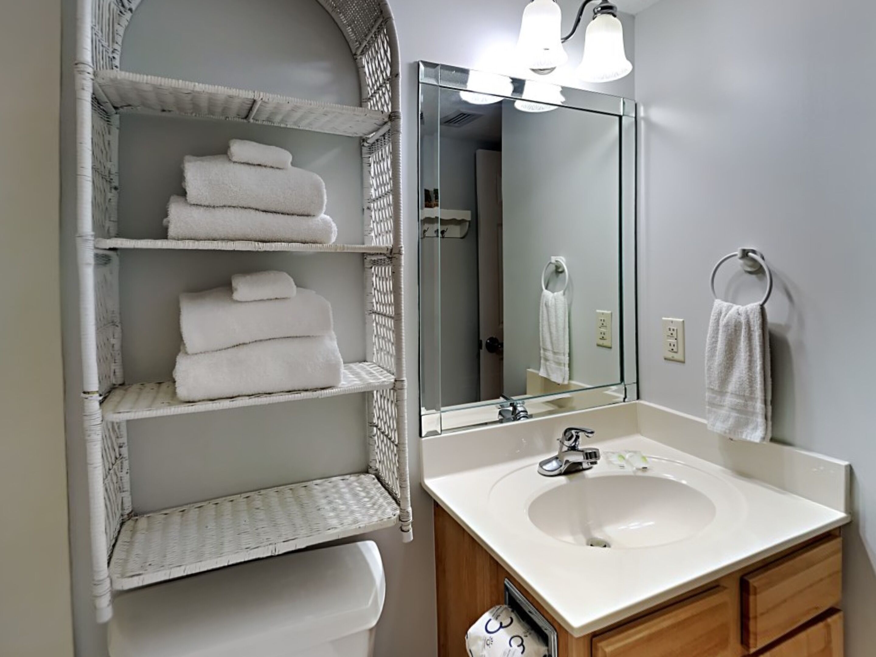 Une salle de bain avec un lavabo blanc, un miroir et des étagères en osier où sont pliées des serviettes.