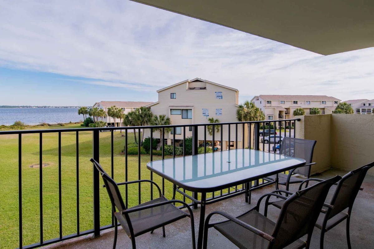 A balcony with a glass table and chairs, overlooking a grassy area and a body of water with buildings in the background.