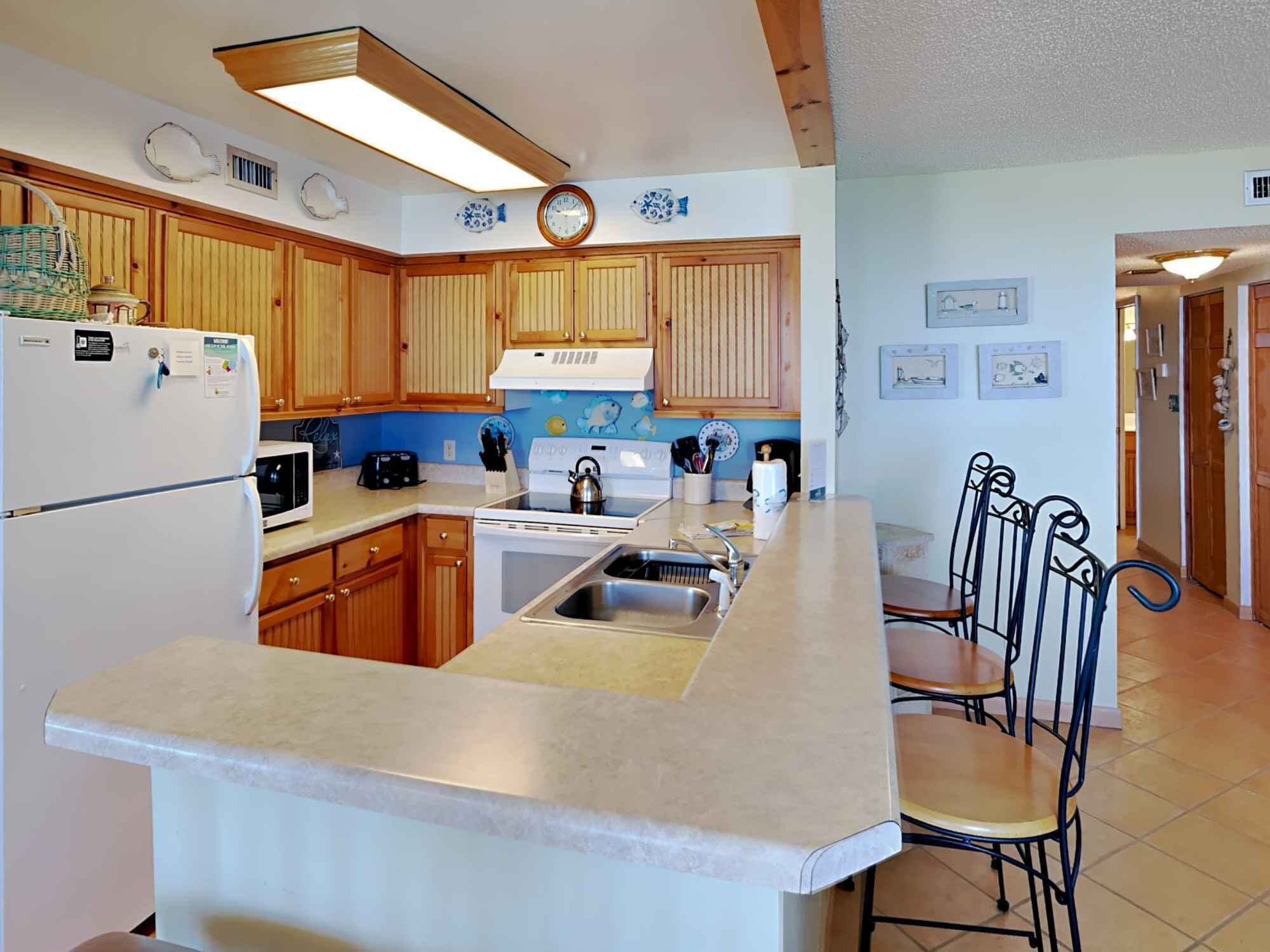 Una cocina con armarios de madera, un refrigerador blanco, una estufa y un lavamanos. Hay dos sillas y una pequeña mesa de comedor.