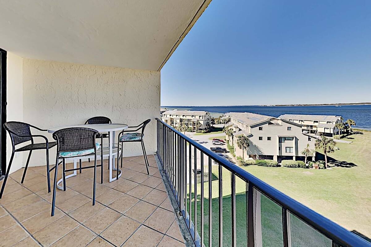 A balcony with a table and chairs, overlooking a beachfront property with buildings and a clear sky.