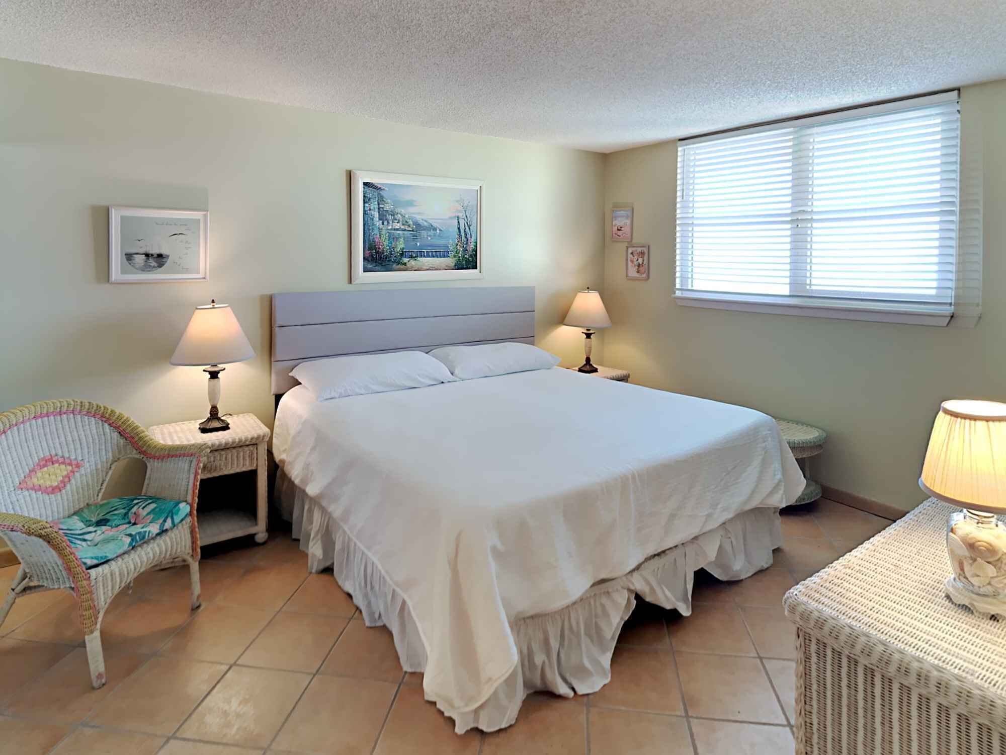 A neatly arranged bedroom with a large bed, two bedside tables with lamps, a chair, and a window with blinds.