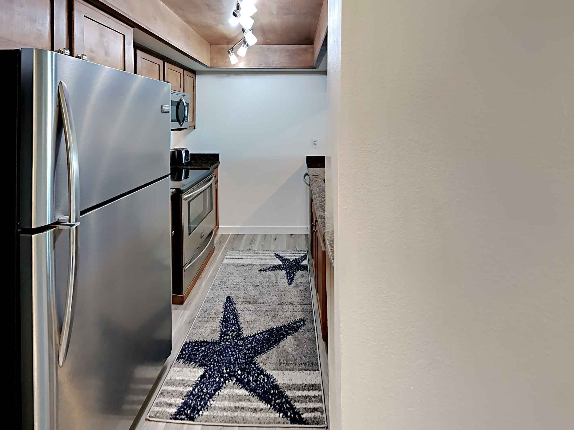 A kitchen with a stainless steel refrigerator, wooden cabinets, a microwave, and a starfish-patterned rug.