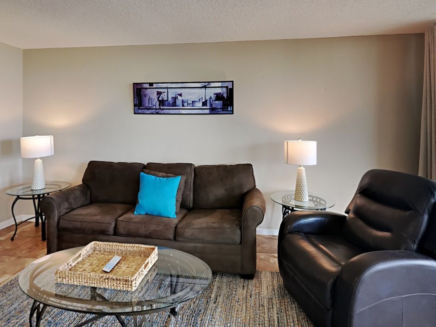 A living room with a brown sofa, a black leather chair, a glass coffee table, and two small side tables with lamps.