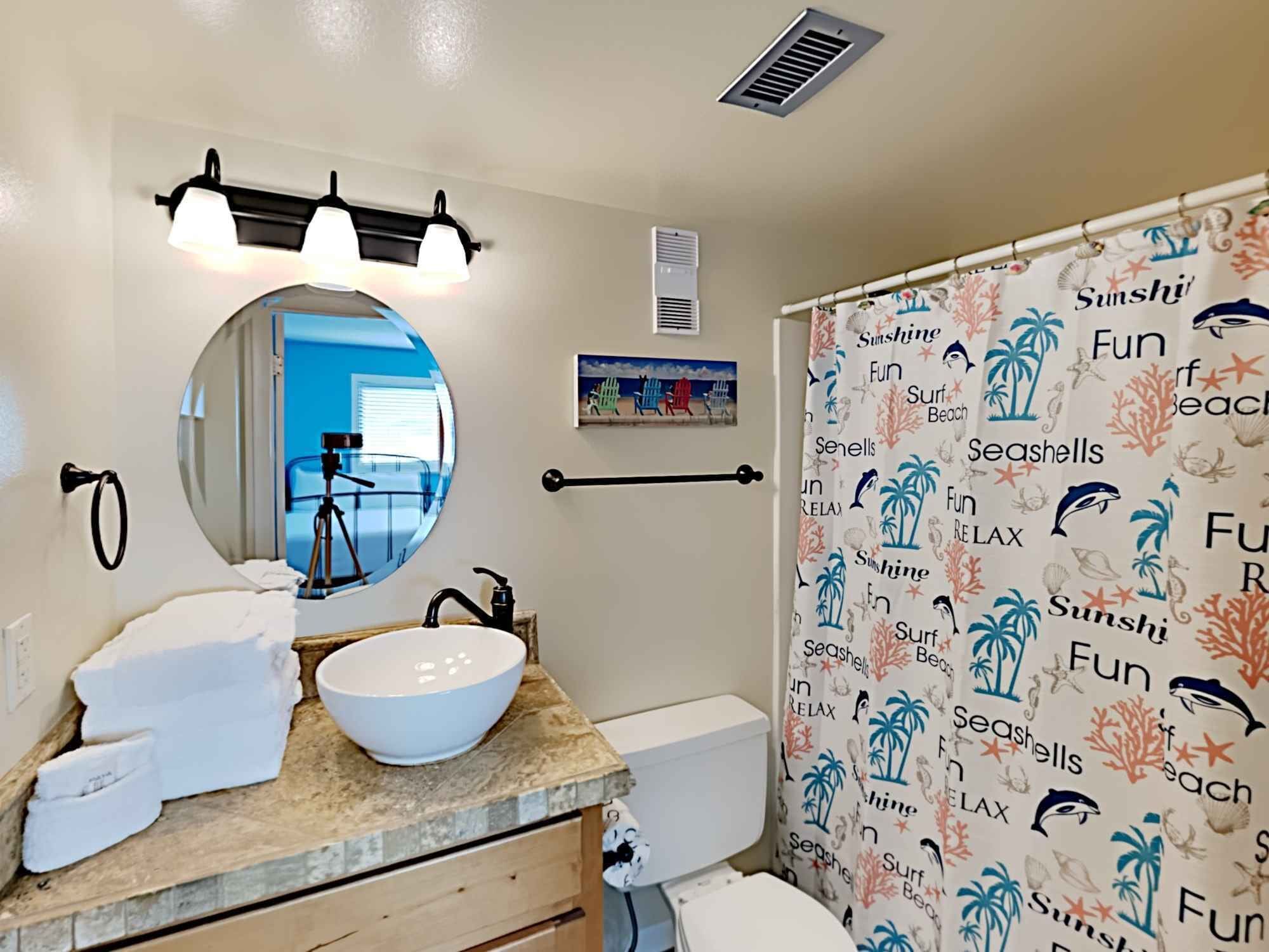 Une salle de bain avec un miroir rond, un lavabo avec un receveur blanc, un porte-serviettes, des toilettes et un rideau de douche sur le thème de la plage.