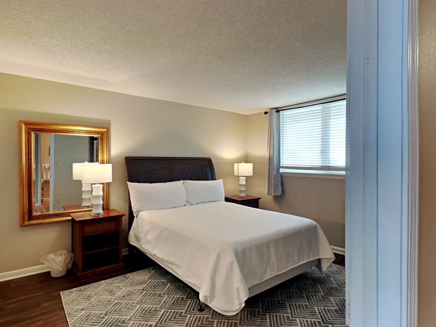 A neatly arranged bedroom with a bed, bedside lamps, a mirror, and a window with blinds.