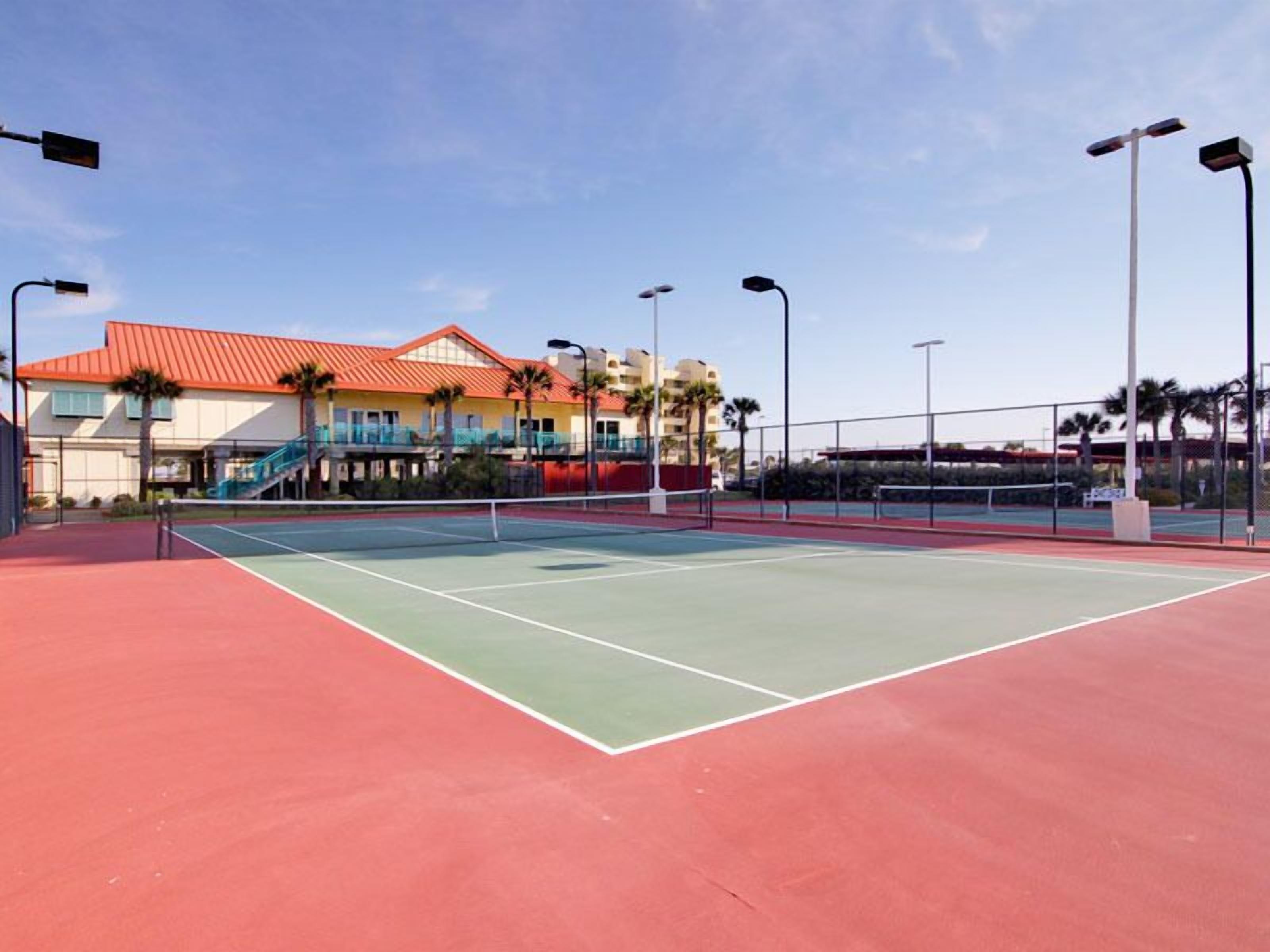 A tennis court with a red and green surface, surrounded by a fence and palm trees.