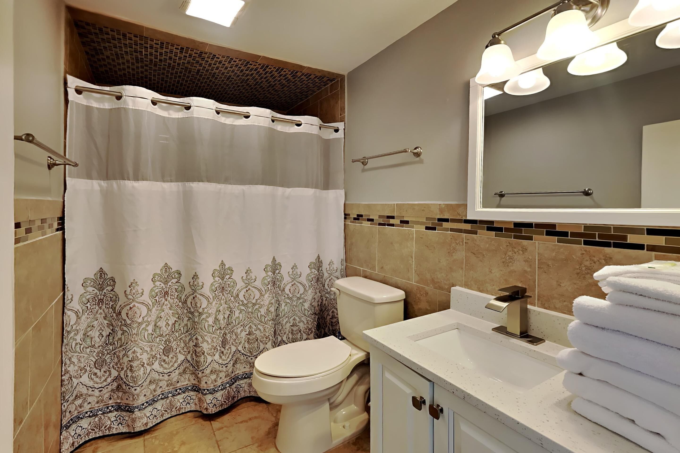 A bathroom with a shower curtain, toilet, sink, and a mirror with lights.