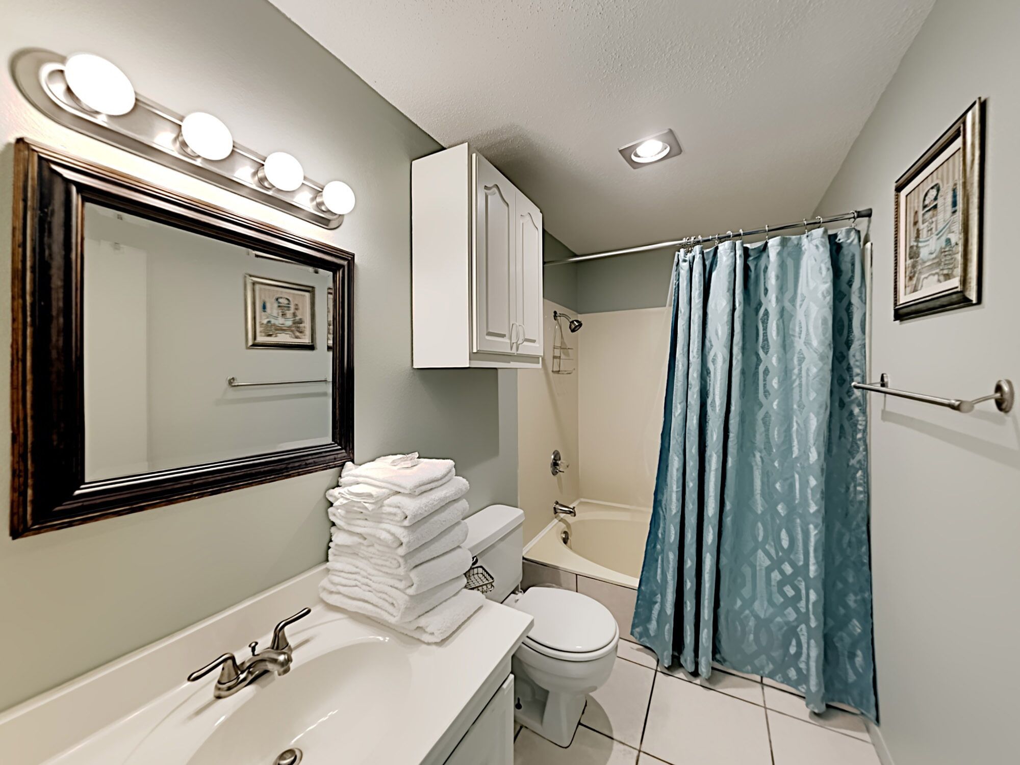A bathroom with a white sink, a mirror with lights, a toilet, a bathtub, and a blue shower curtain.
