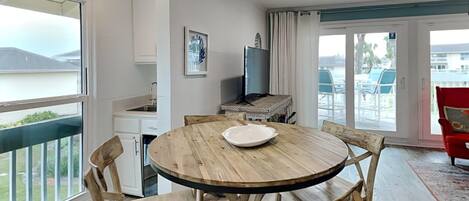 A dining area with a round wooden table and chairs, a TV on a stand, and a view of the outdoors through large windows.