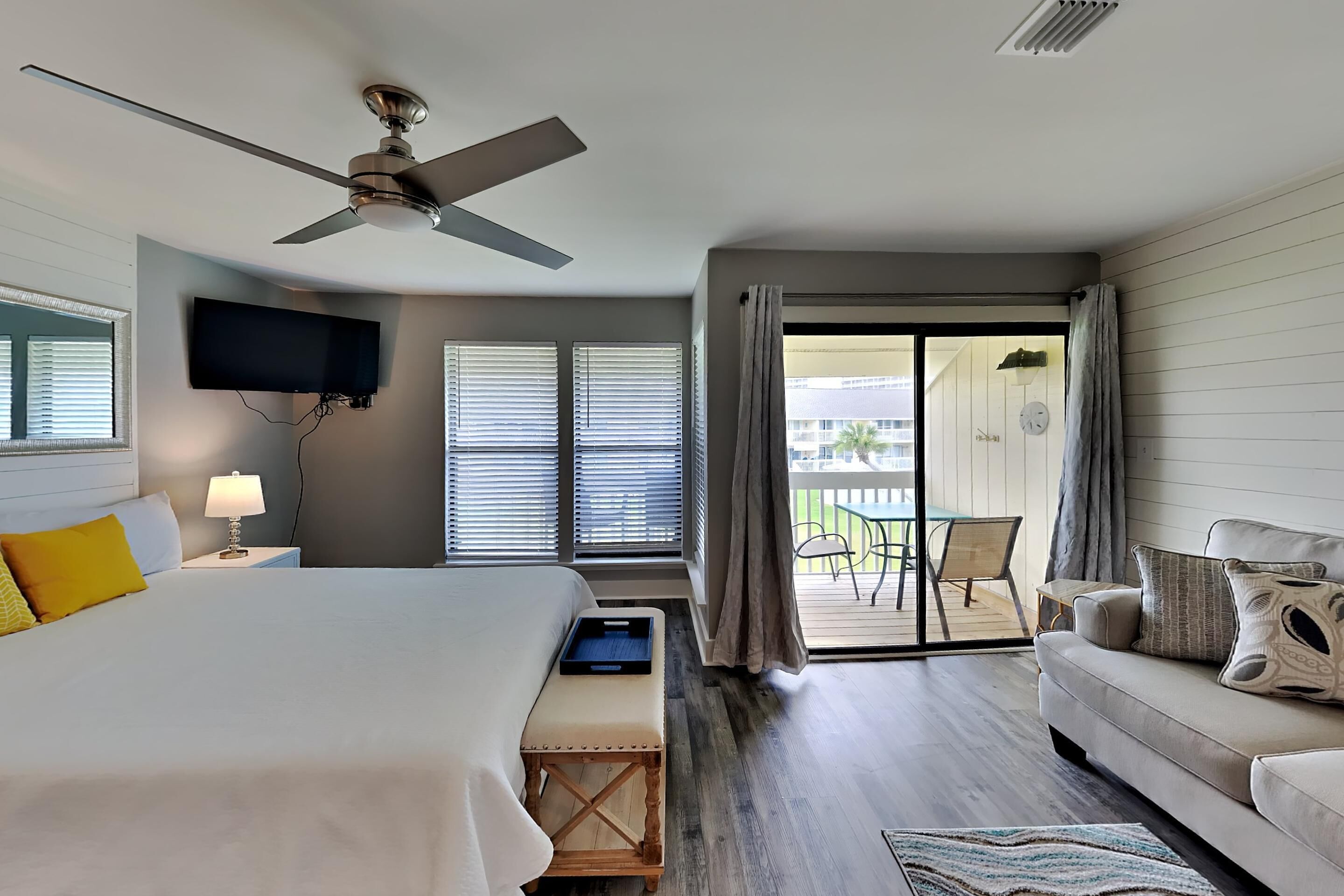 A hotel room with a bed, a sofa, a TV, and a balcony with outdoor furniture.