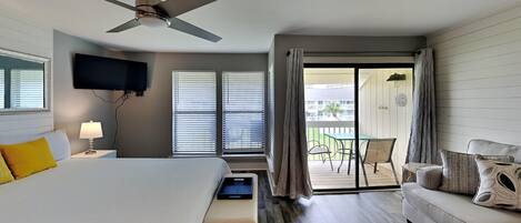 A hotel room with a bed, a sofa, a TV, and a balcony with outdoor furniture.