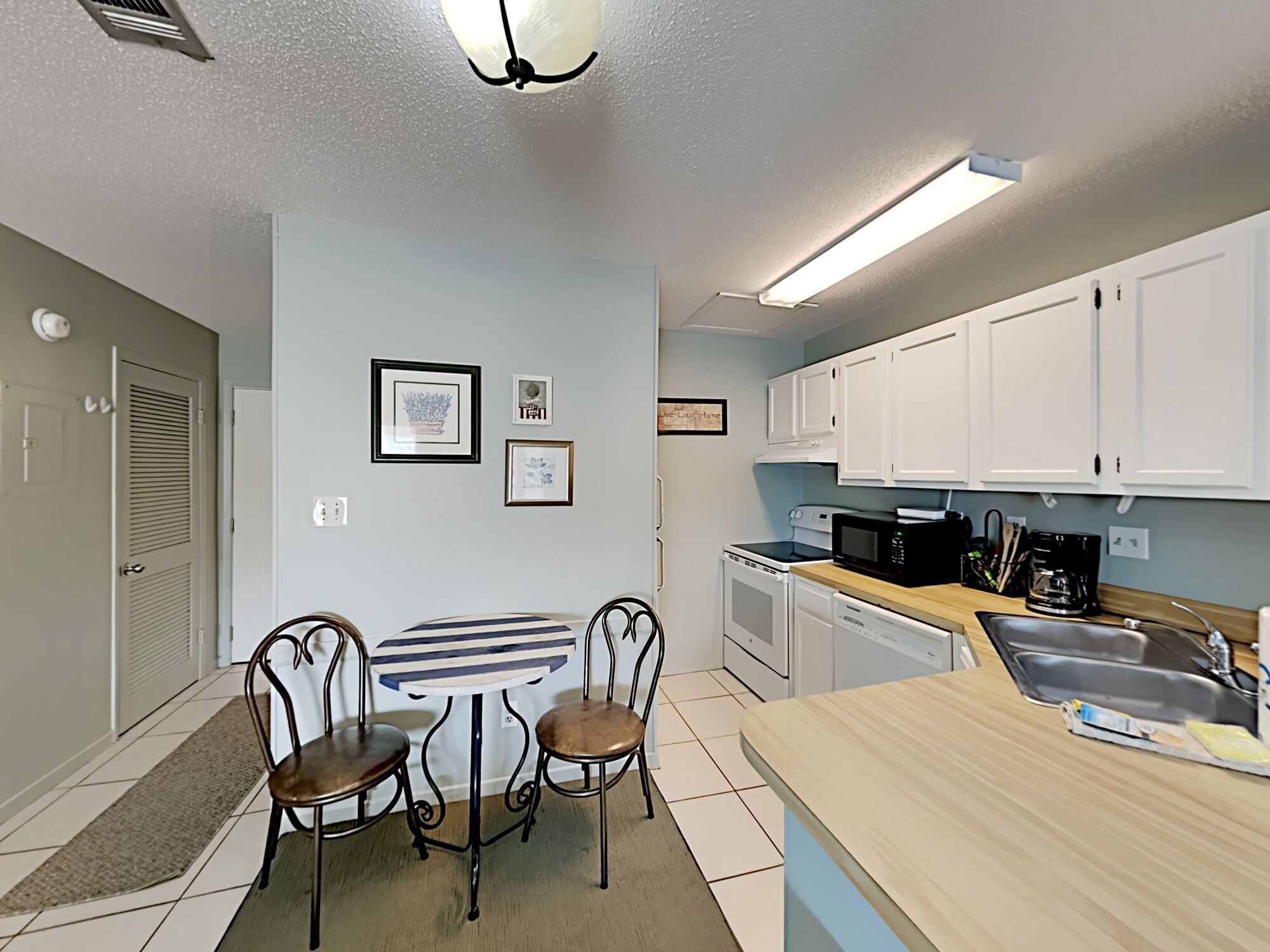A compact kitchen with white cabinets, a microwave, and a sink. There is a striped table with two chairs and a small rug.