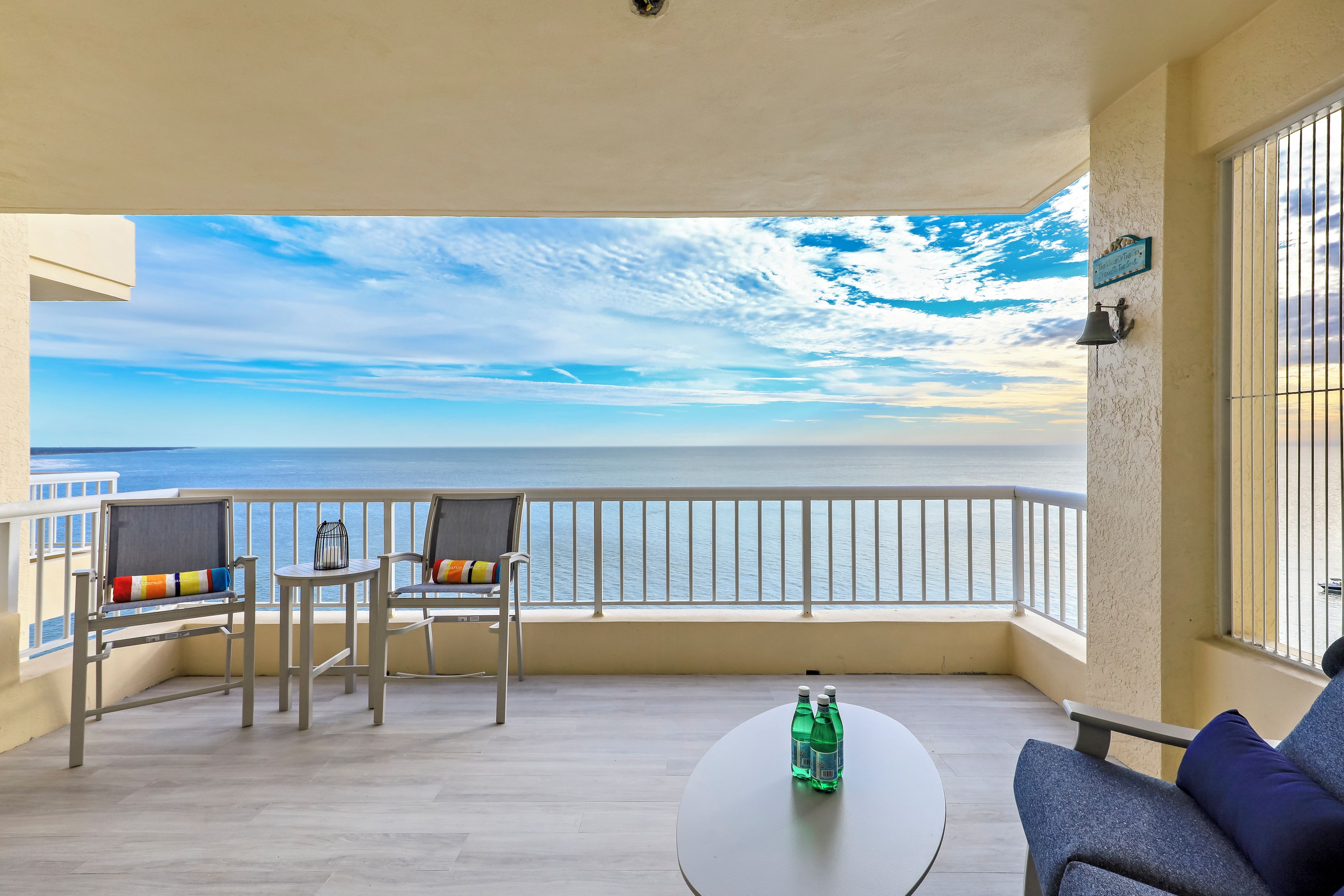 Un balcón con vistas al mar, una mesa redonda con botellas y dos sillas con cojines de colores.