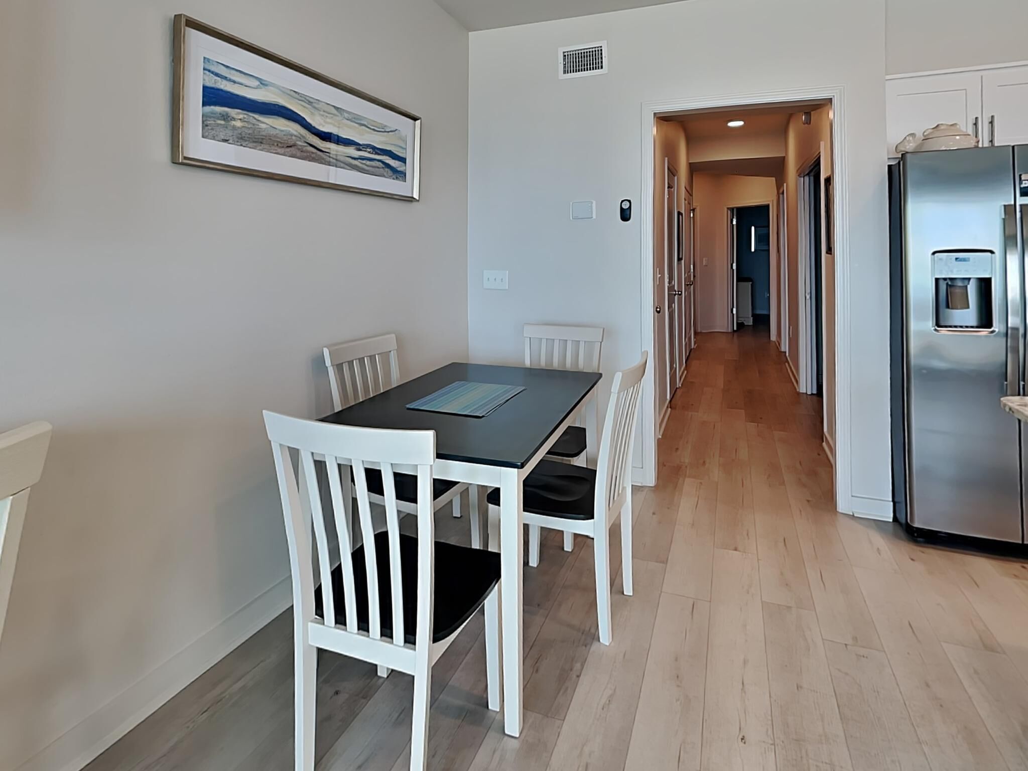 Uma sala de jantar com mesa e cadeiras, uma cozinha com frigorífico, e um corredor que dá para outro quarto.